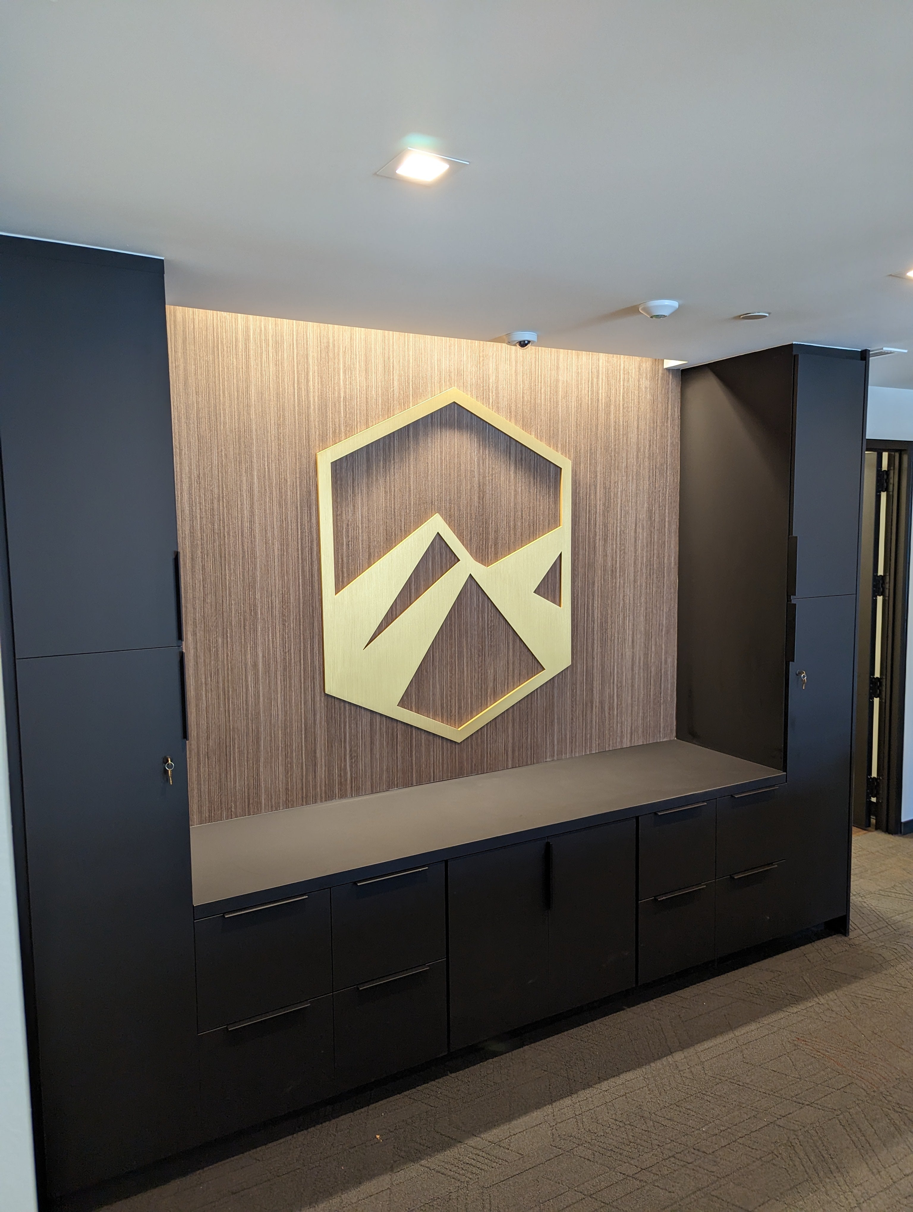 An elegant indoor lobby sign for a company in Denver, Colorado. The sign consists of a minimalist geometric mountain logo in gold, mounted on a textured wood-grain wall. The surrounding area features sleek, black cabinetry beneath the logo, creating a modern and polished corporate interior.
