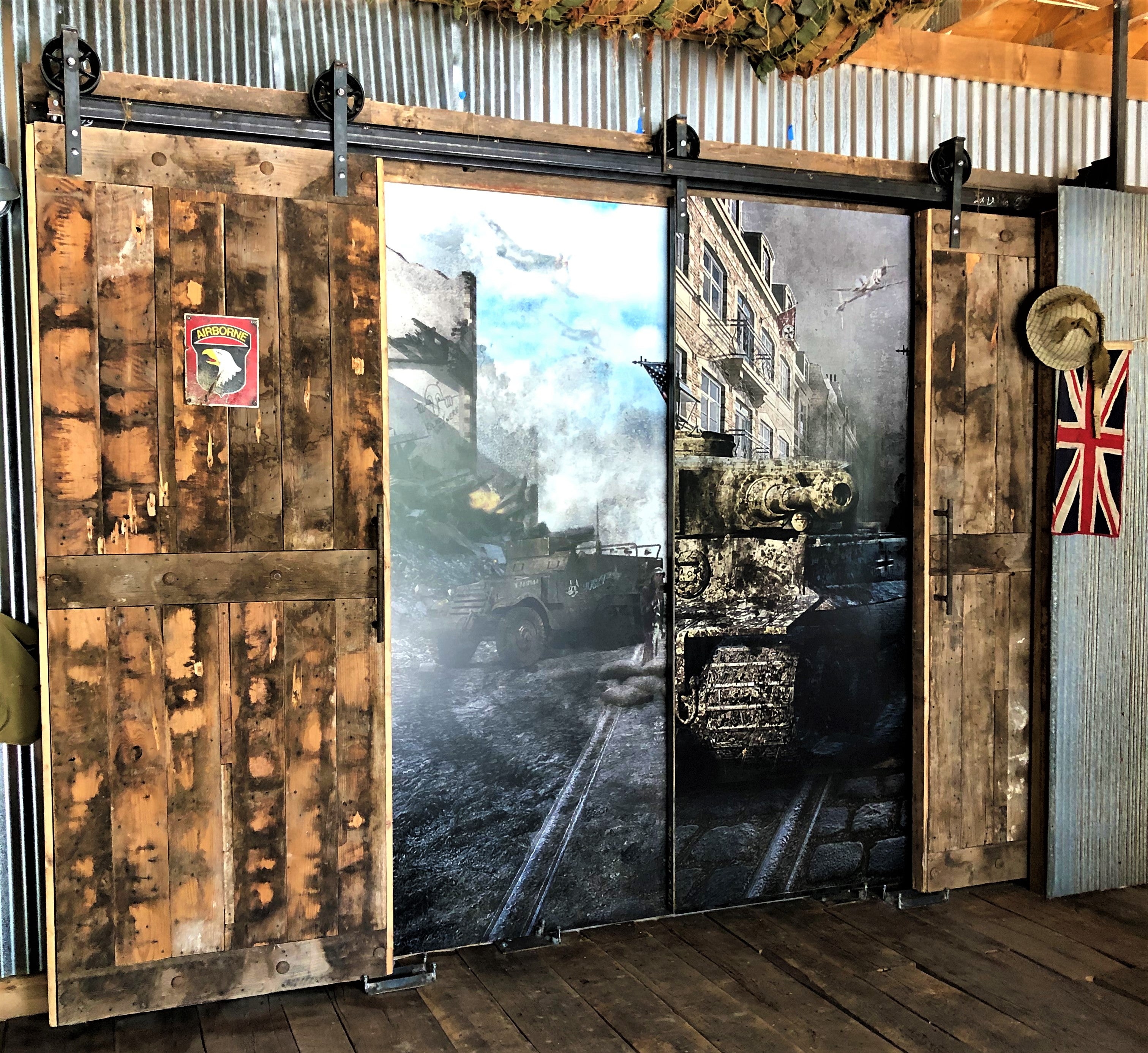 A rustic, industrial-themed interior vinyl sign featuring sliding barn doors with a World War II tank and street scene graphic. The left side has a worn wooden finish with an "Airborne" patch, while the center and right panels display a military tank amidst urban ruins. A British flag and hat hang on the side.