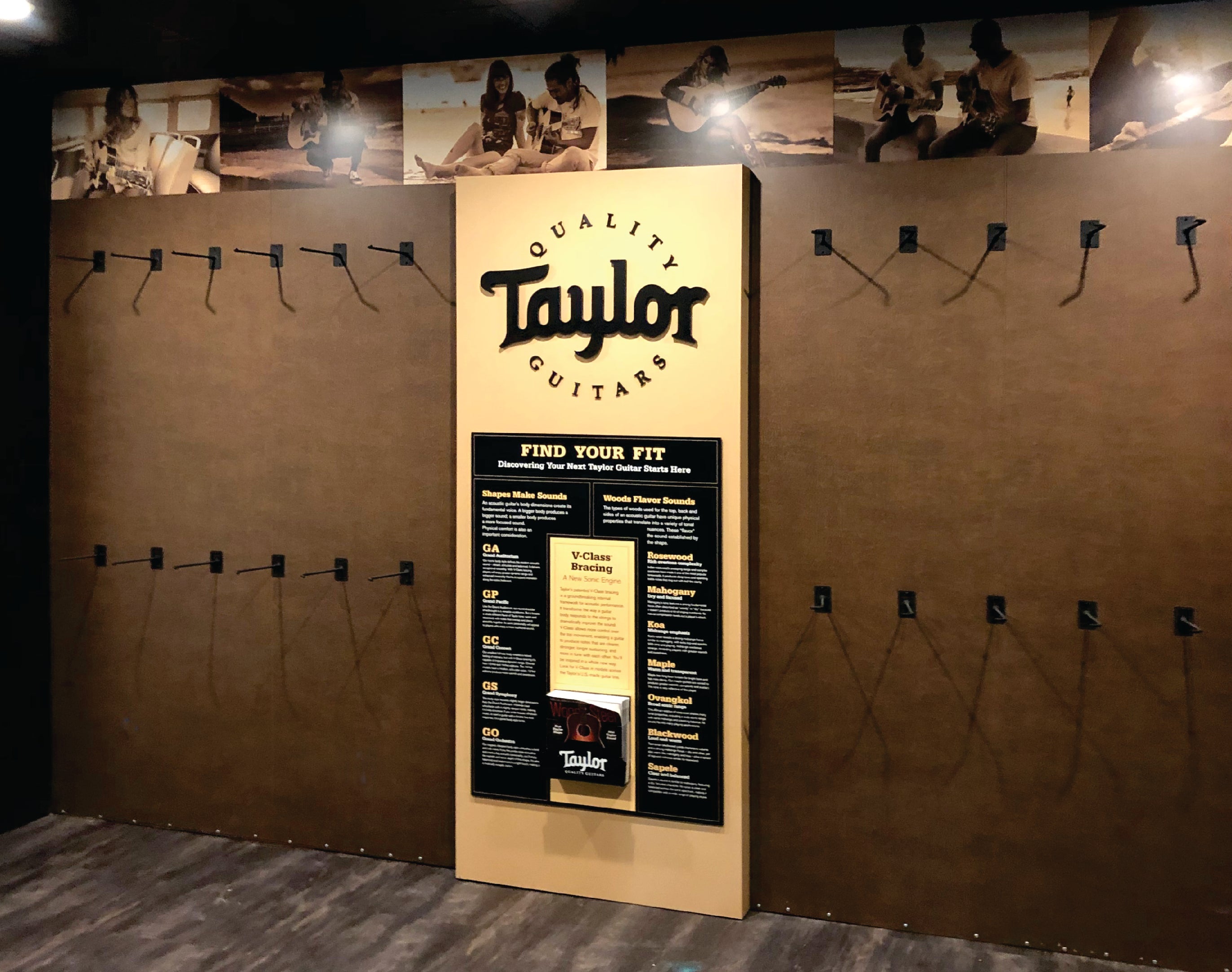 An interior wall sign for Taylor Quality Guitars in Broomfield, Colorado. The display features a wood-paneled wall with the Taylor Guitars logo prominently displayed at the top in bold, black lettering. Below the logo, a section titled "FIND YOUR FIT" provides information on guitar models, bracing, and wood types. The display is framed by empty guitar hooks, suggesting it is part of a showroom or retail space for showcasing Taylor guitars.