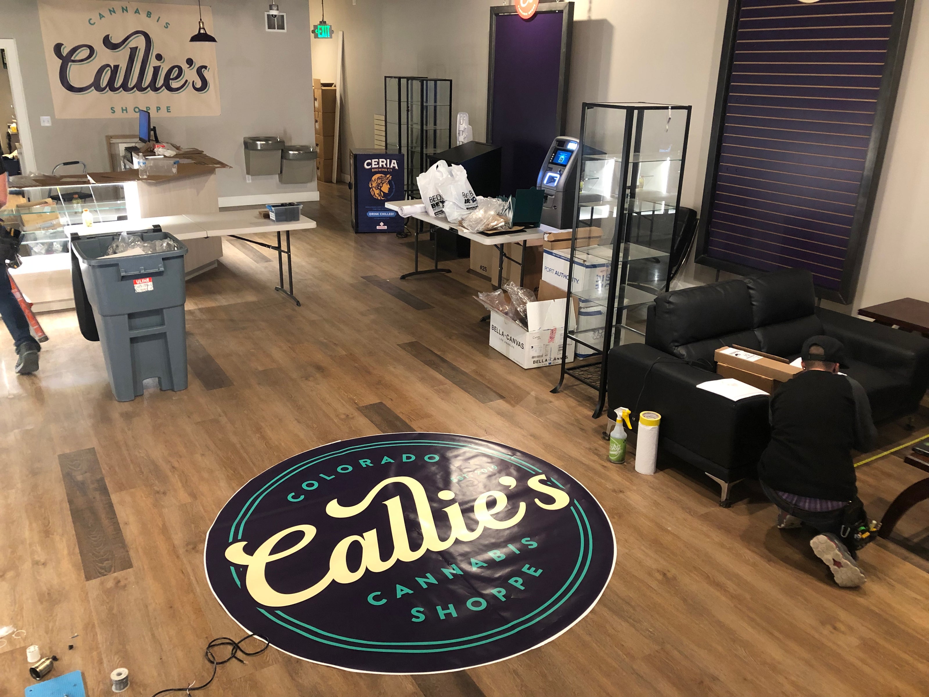 A custom circular floor wrap placed in the center of the shop's wooden floor, featuring "Colorado Callie’s Cannabis Shoppe" in bold, colorful typography. The shop space is being organized with equipment near the floor wrap installation. This project is for Callie's Cannabis Shoppe in Northglenn, Colorado.