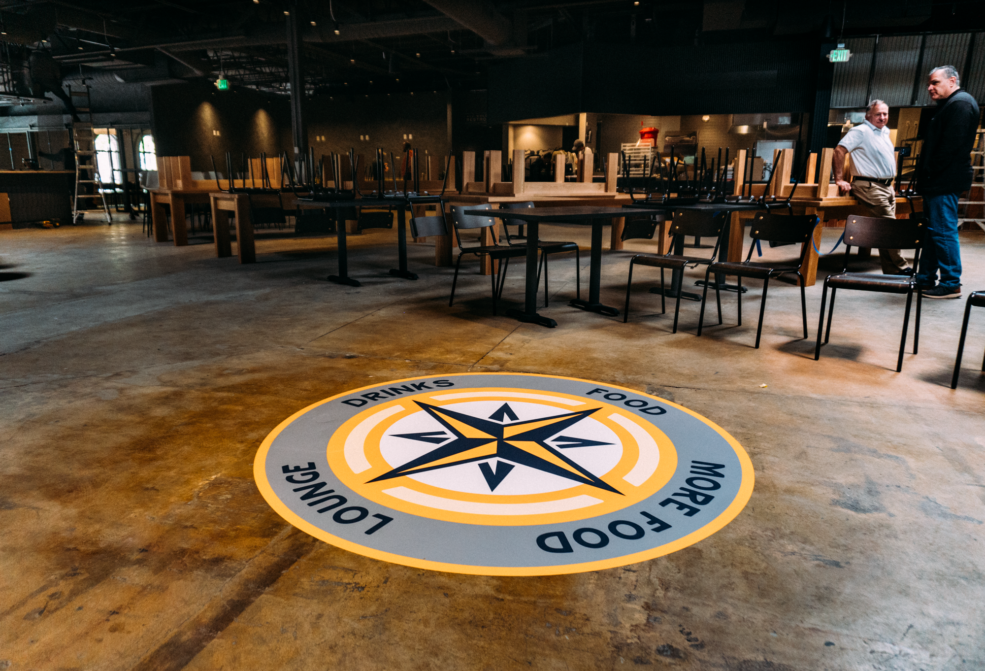 Another view of the interior floor wrap at Parkway Food Hall in Longmont, Colorado, highlighting the vibrant compass design and its placement in the bustling food hall setting, with seating and food stations visible in the background.