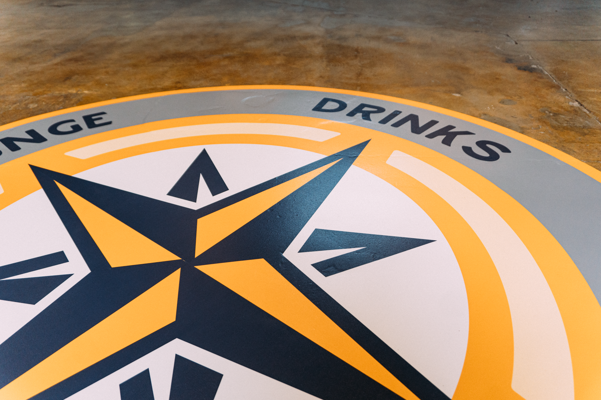 Another angle of the graphic floor wrap at Parkway Food Hall in Longmont, Colorado, focusing on the detailed compass design that dominates the visual, surrounded by the food hall's interior setting with scattered furniture in the background.