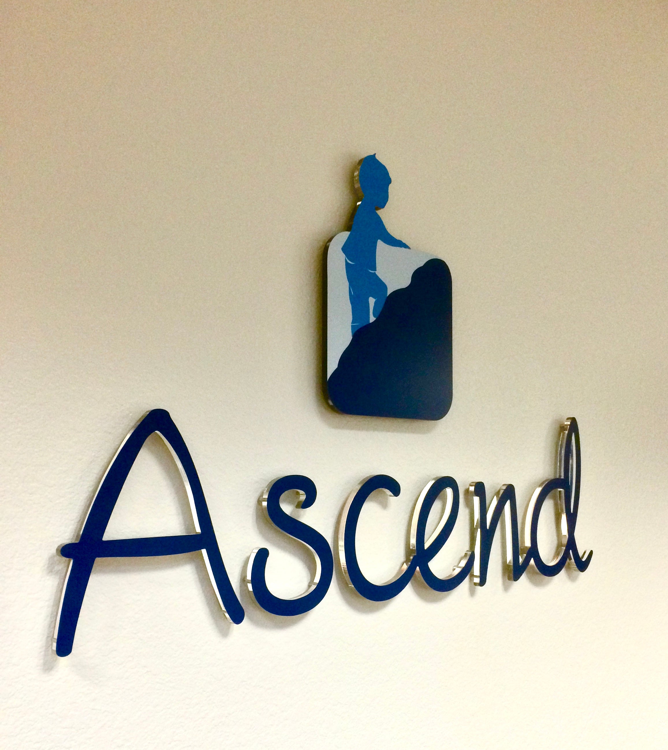 A wall-mounted sign for Ascend in Boulder, Colorado. Featuring acrylic lettering for their name and a metal cutout for their logo of a child climbing a hill.