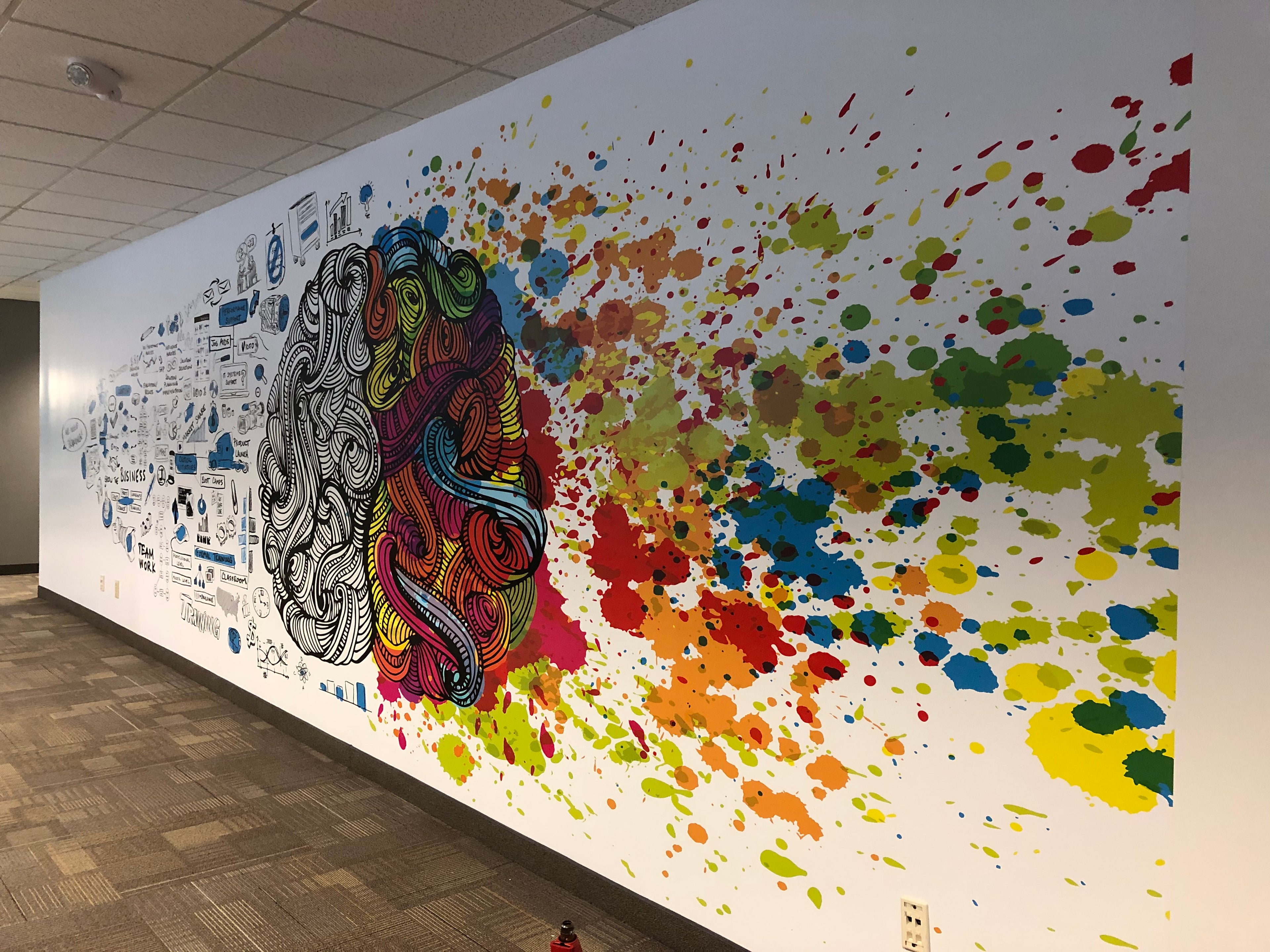 A colorful, creative wall wrap inside the Volvo Mack building in Atlanta, Georgia. The design shows an artistic representation of a brain, with the left half filled with detailed illustrations and words, while the right half explodes into vibrant splashes of color. The wrap spans a large portion of the wall.