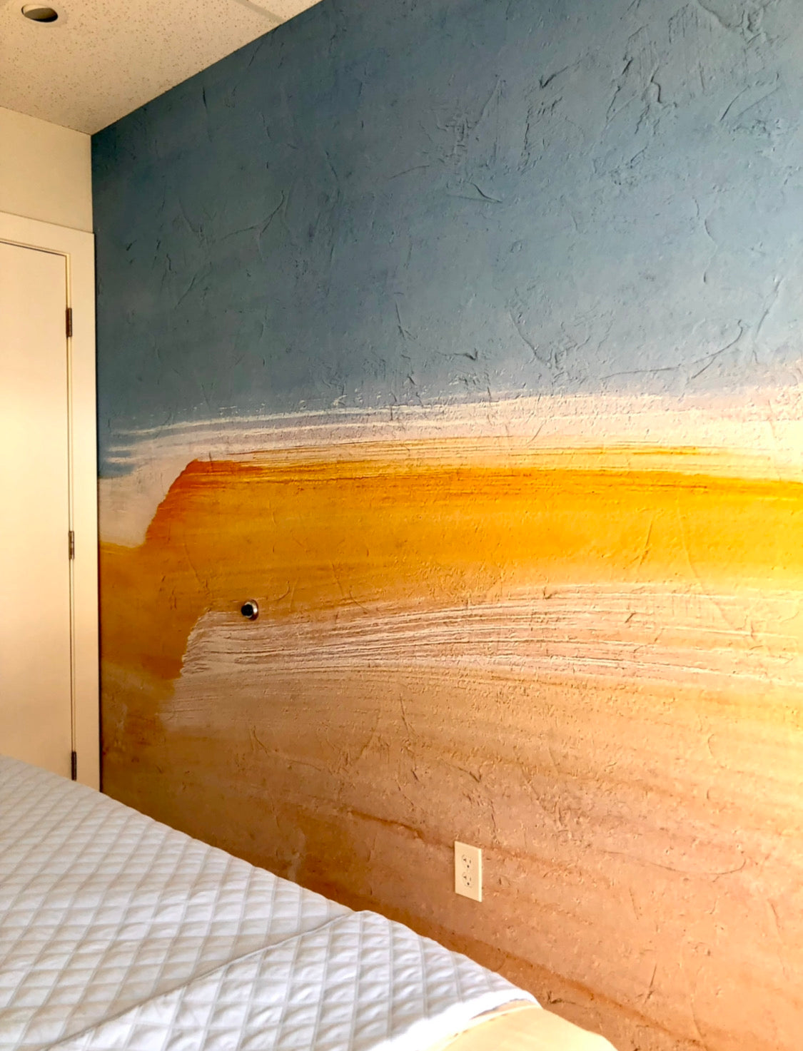 A large vinyl wall wrap inside a room, featuring a soft blend of blue, white, and earthy tones. The design mimics a serene horizon line, creating a calm and soothing atmosphere. The textured surface adds a dimensional effect to the wrap.