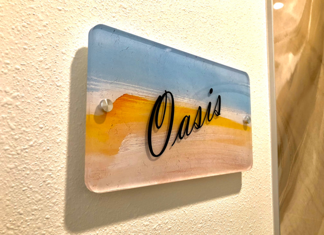 A small wall-mounted acrylic wayfinding sign that reads "Oasis" in elegant black script. The background features a warm, gradient blend of soft blues, oranges, and yellows, giving the sign a soothing, sunset-like appearance. The sign is attached to a textured wall using silver standoffs.