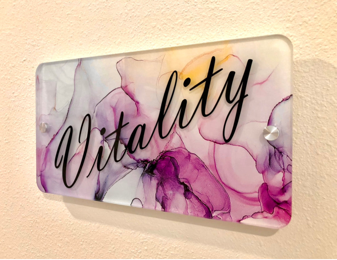 A stylish wayfinding sign featuring the word "Vitality" in black script on a vibrant purple and pink abstract background. This sign also includes metal standoffs, part of a wayfinding system at The Body Bar in Lafayette, Colorado.