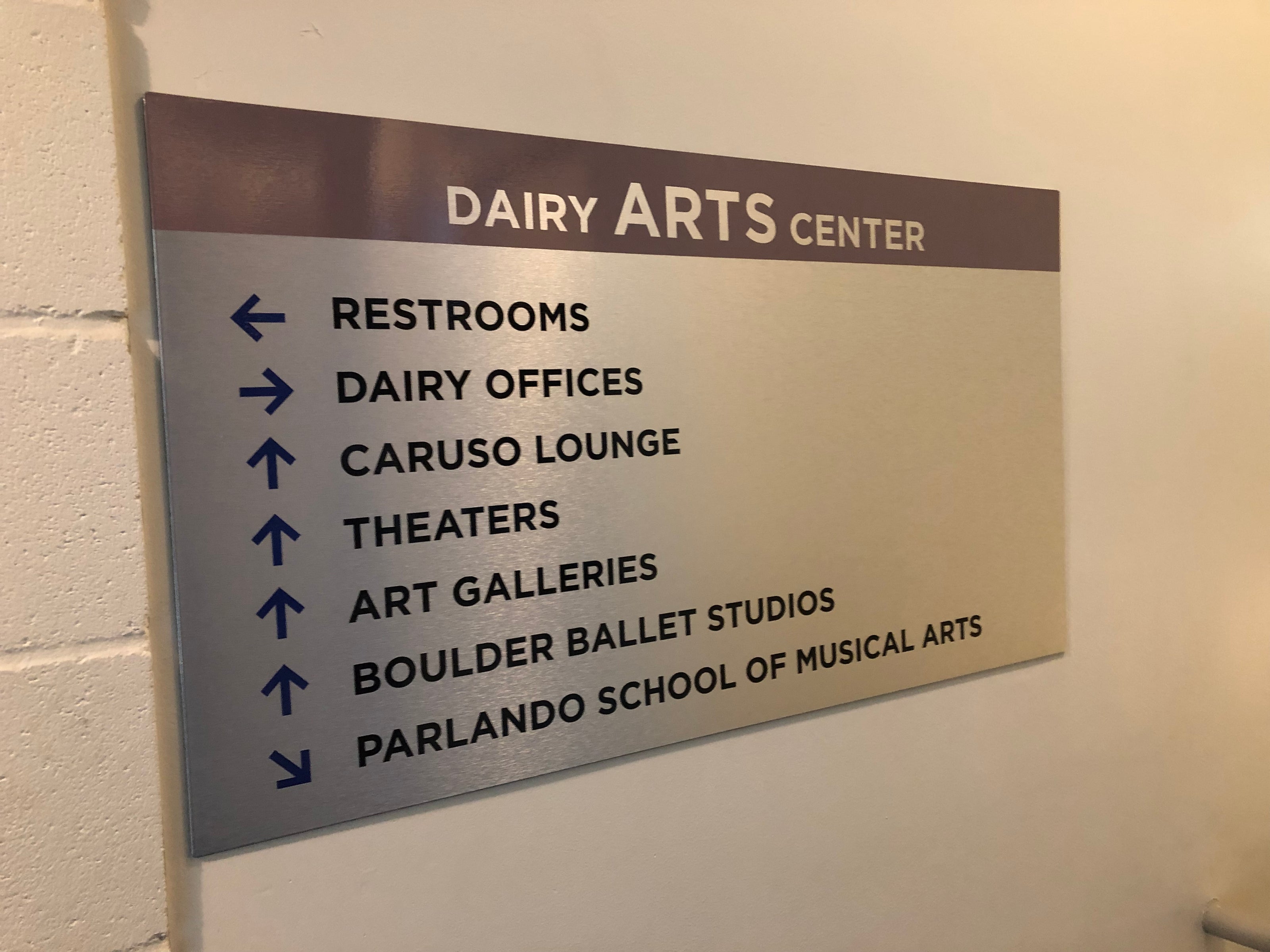 A metal wayfinding sign with silver and purple colors, directing visitors to various parts of the Dairy Arts Center, such as restrooms, Dairy offices, Caruso Lounge, theaters, art galleries, Boulder Ballet Studios, and Parlando School of Musical Arts. This ADA-compliant sign is located at the Dairy Arts Center in Boulder, Colorado.