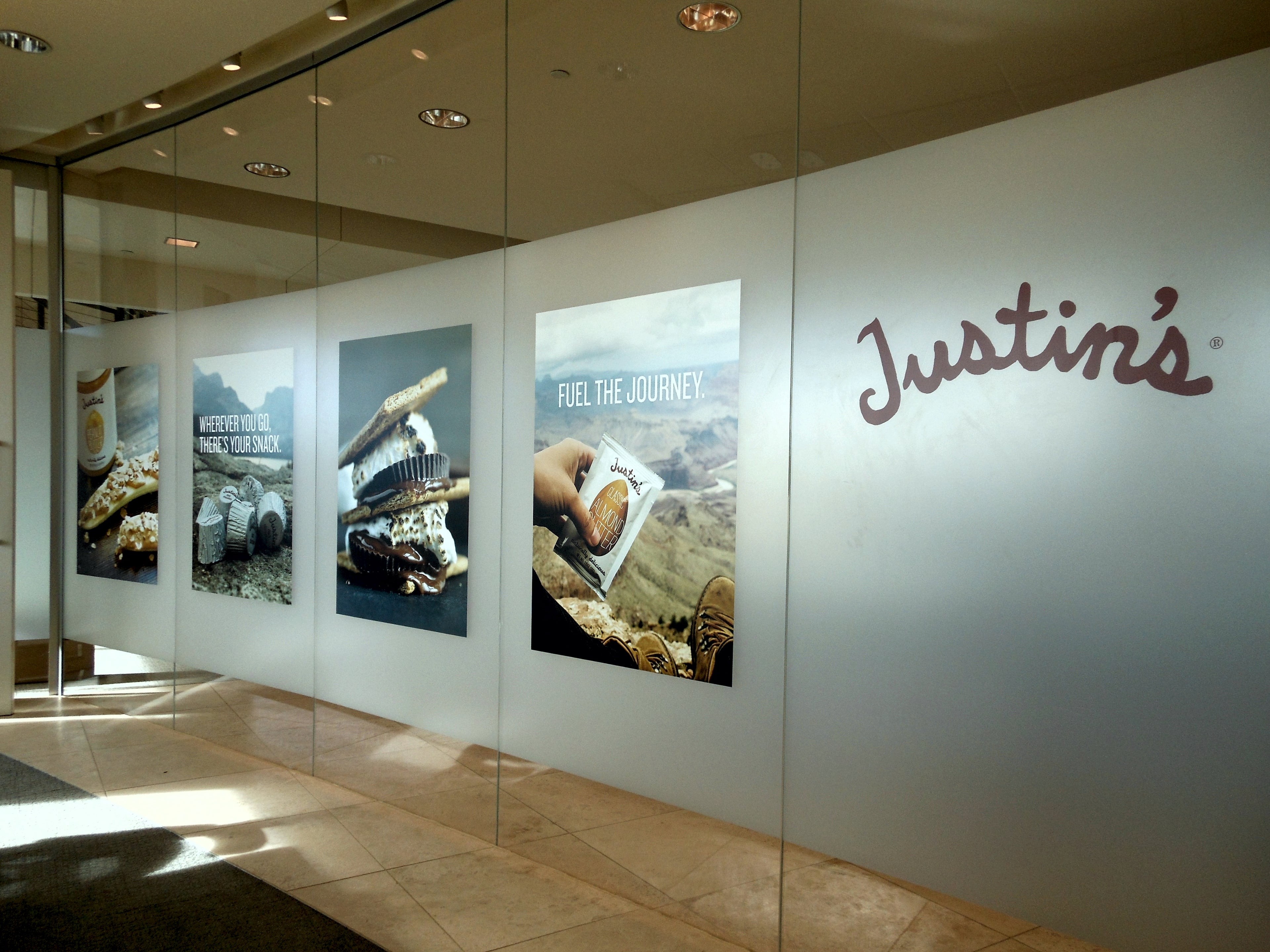 An indoor installation featuring printed frosted glass vinyl wrap created for Justin's Nut Butter in Boulder, Colorado. The design showcases several product images and lifestyle visuals, including peanut butter cups and snacks, along with phrases like "Fuel the Journey" and "Wherever You Go, There's Your Snack." The Justin's logo is prominently displayed on the right side.