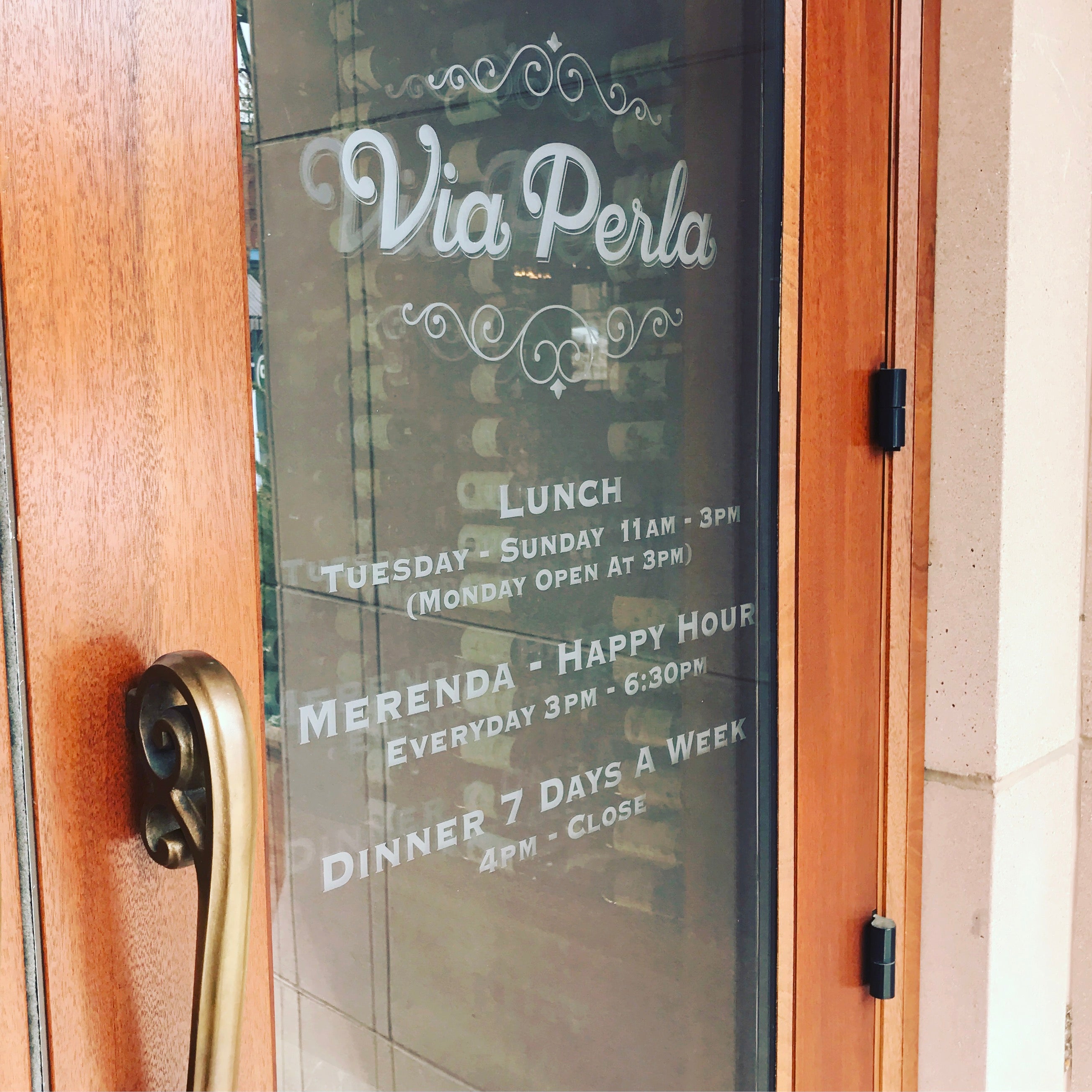 A close-up of an elegant window wrap at Via Perla in Boulder, Colorado. The window displays the restaurant's name "Via Perla" with hours of operation in a classic white script, framed by a wooden door for a sophisticated, welcoming entrance.
