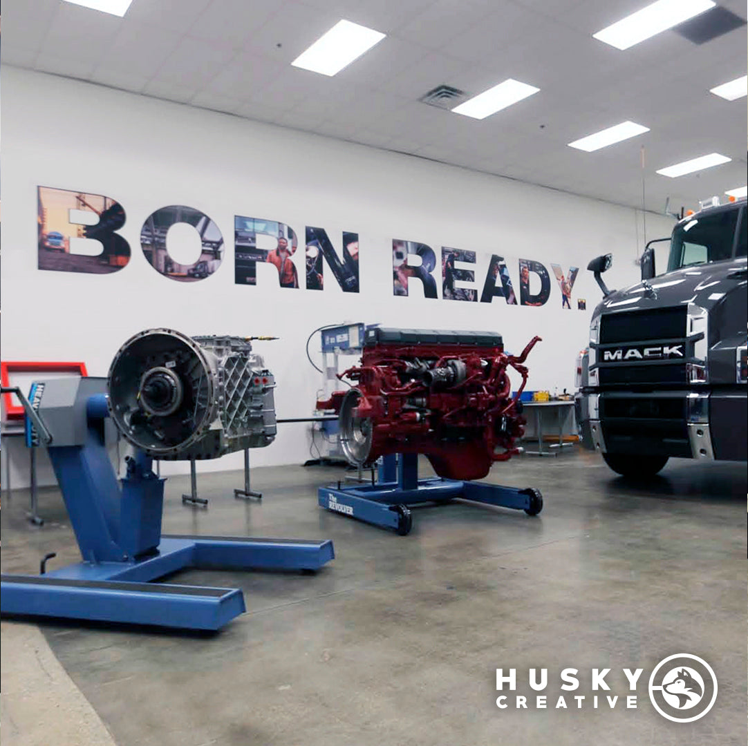 Vinyl lettering reading "Born Ready." on a wall at Volvo Mack in Atlanta, Georgia with a Mack truck and an engine in front of it.