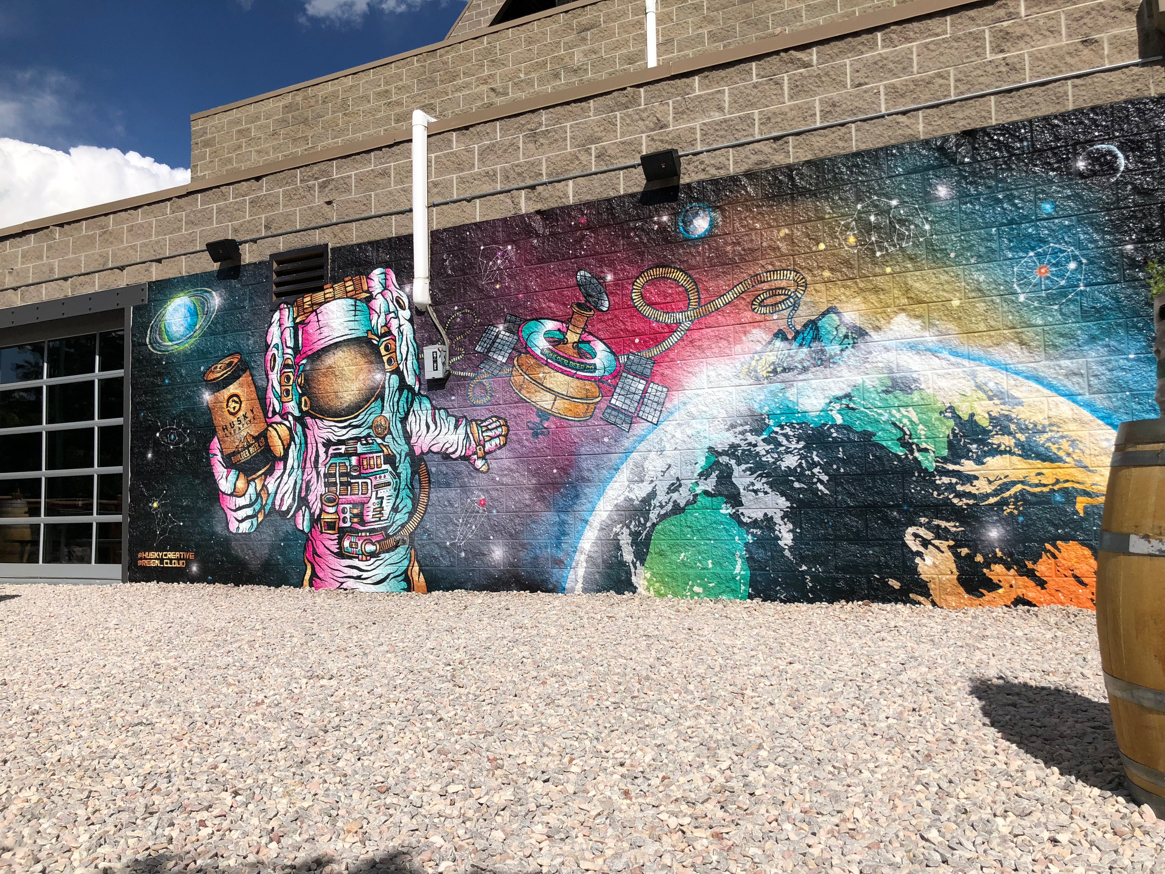 A vibrant and eye-catching building wrap depicting an astronaut holding a beer can, floating in space with Earth and satellite imagery in the background. This colorful mural adorns the exterior of a brewery in Boulder, Colorado, creating a unique and engaging visual for passerby.