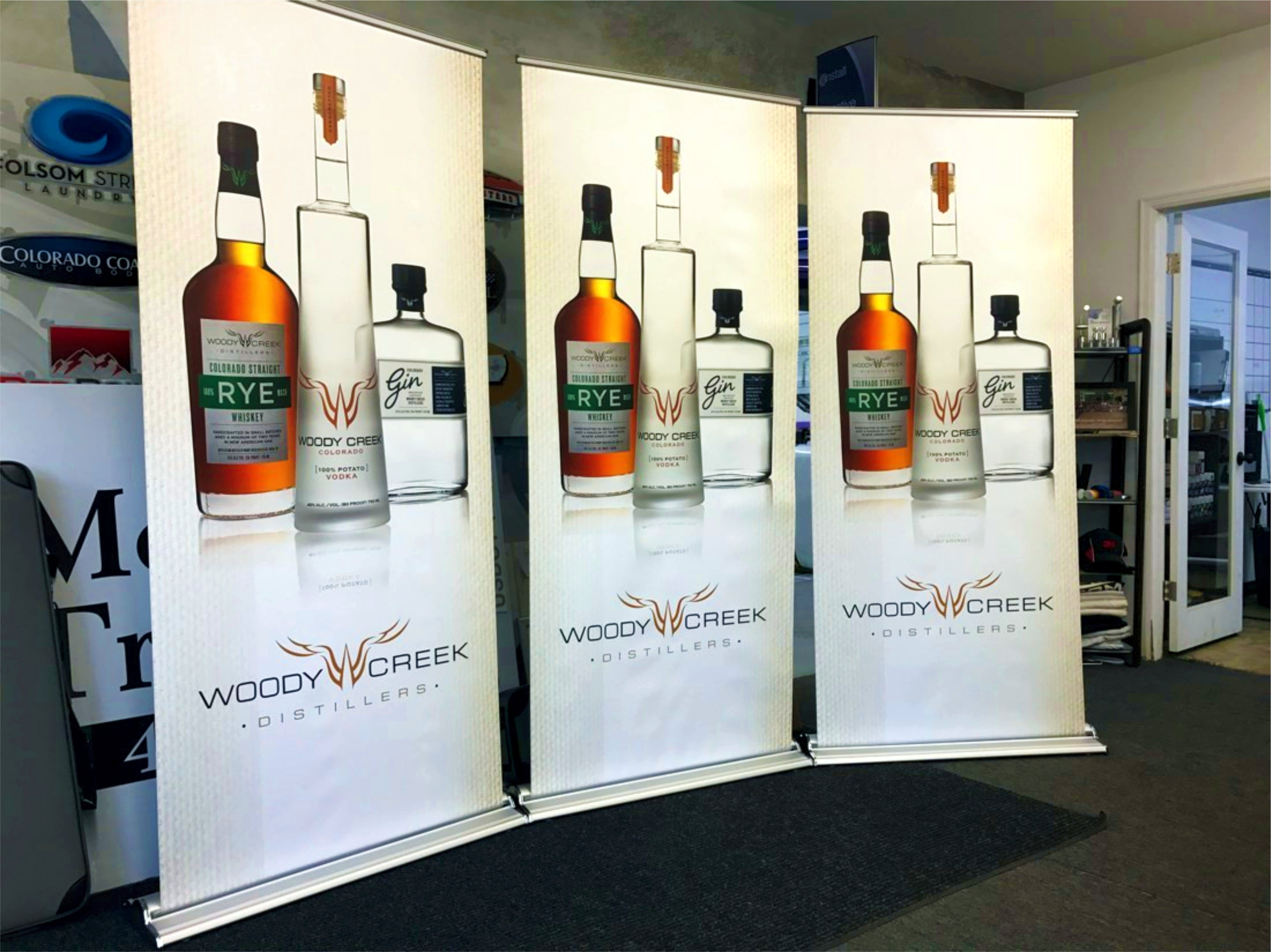 A set of three retractable banners featuring large, high-quality images of whiskey, vodka, and gin bottles from Woody Creek Distillers, based in Basalt, Colorado. The bottles are centered on a white background with the distillery's logo below, creating a sleek and professional look.