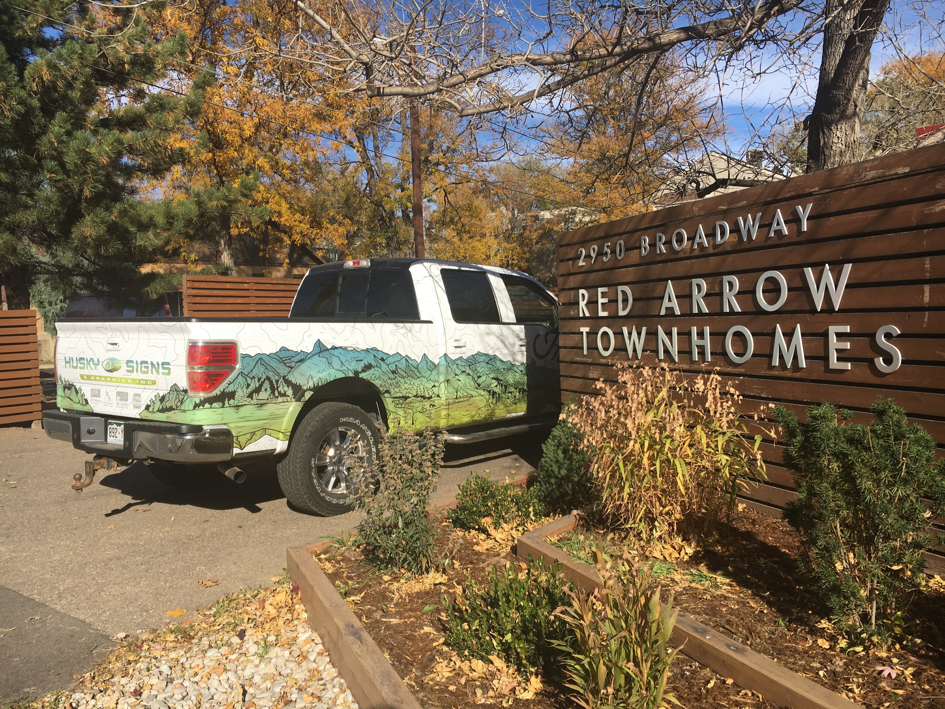 A pickup truck with a custom vehicle wrap featuring a green landscape scene, parked next to the monument sign for Red Arrow Townhomes in Boulder, Colorado. The truck wrap shows a stylized mountain range with the Husky Signs logo, complementing the natural wood and greenery surrounding the location.