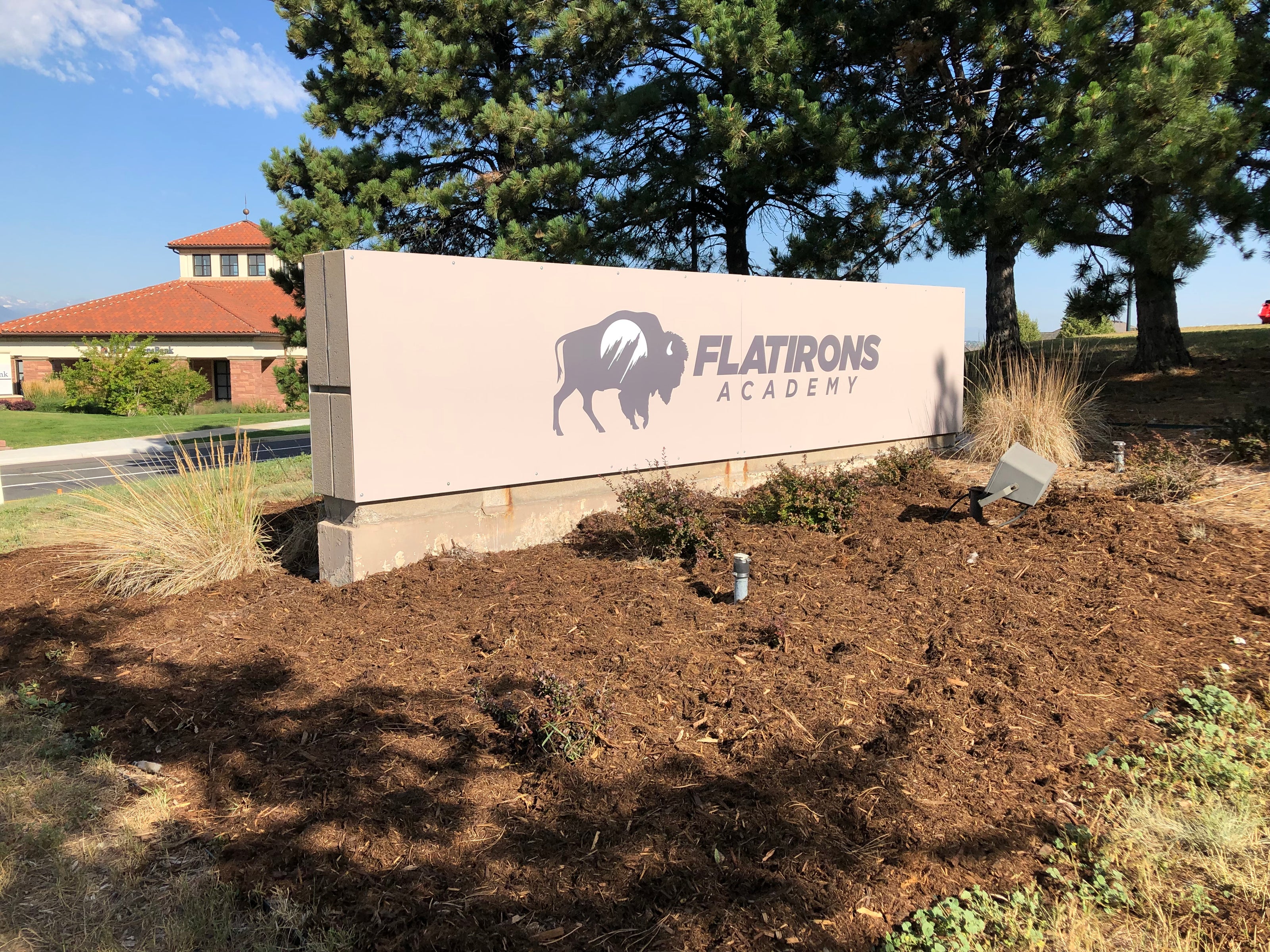 A wide, beige, and rectangular monument sign for Flatirons Academy, featuring a bison logo in gray with the name in bold letters. The sign is placed in a landscaped area with mulch and plants in front, and tall trees and a red-roofed building in the background, located on the CU Boulder campus in Boulder, Colorado.