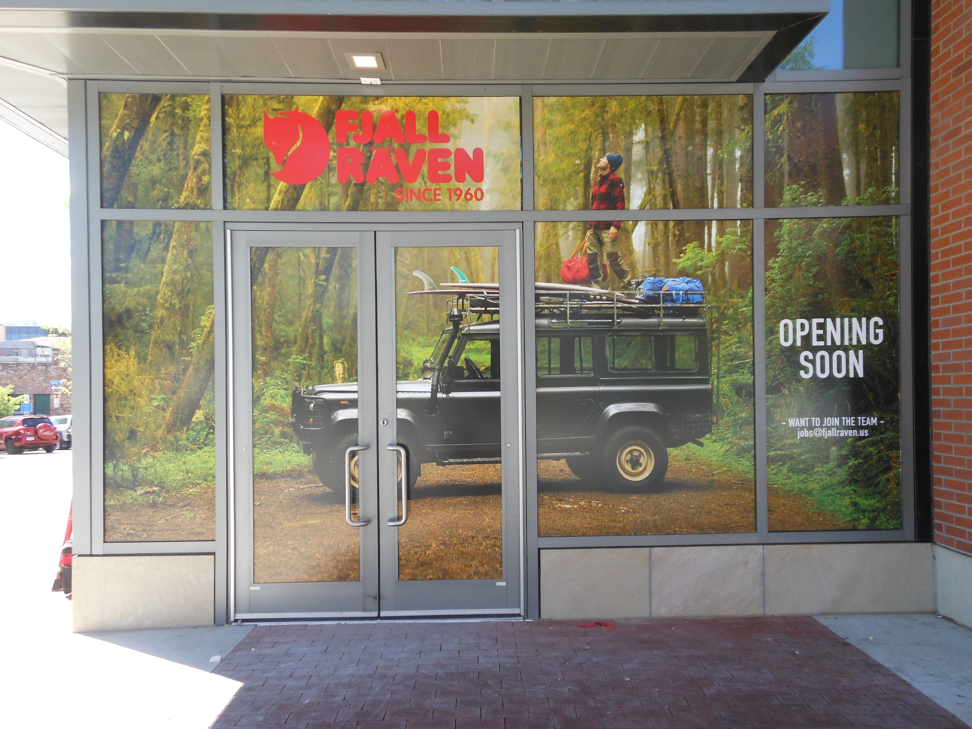 A storefront window wrap installed for Fjall Raven at their Boulder, Colorado location. The wrap features a scenic forest backdrop with a black Land Rover and an adventurer standing on top. The Fjall Raven logo is prominently displayed in red, along with the text "Opening Soon" on the right side.