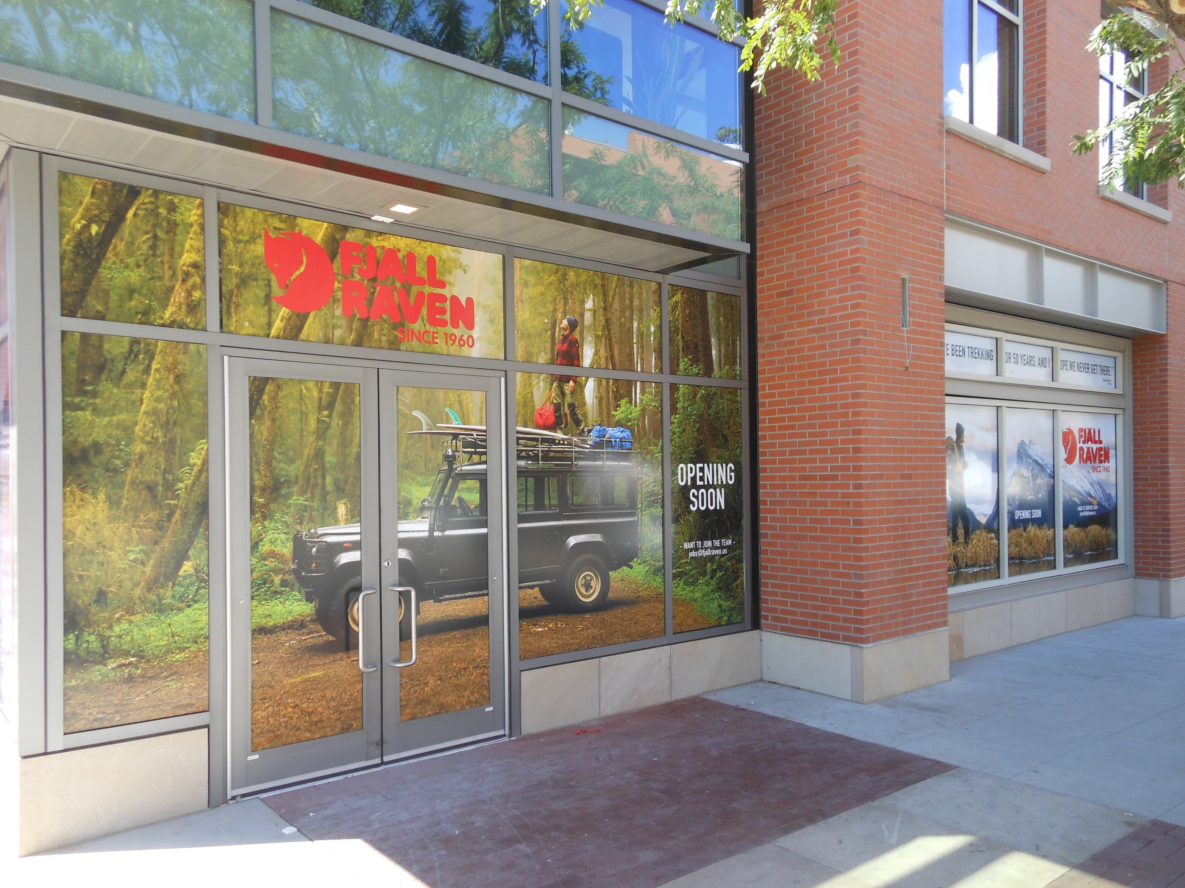 The exterior of a Fjall Raven store in Boulder, Colorado, showcasing vibrant outdoor-themed window wraps with images of nature, mountains, and trekking gear. The main entrance is covered with a large wrap featuring a forest setting and a Land Rover, enhancing the store's rugged, adventurous brand image.