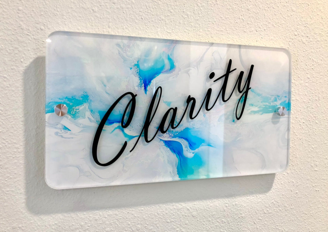 A sleek, modern wall sign with the word "Clarity" in elegant black script on a white and blue abstract background. The sign is mounted with metal standoffs for a floating effect, located at The Body Bar in Lafayette, Colorado.