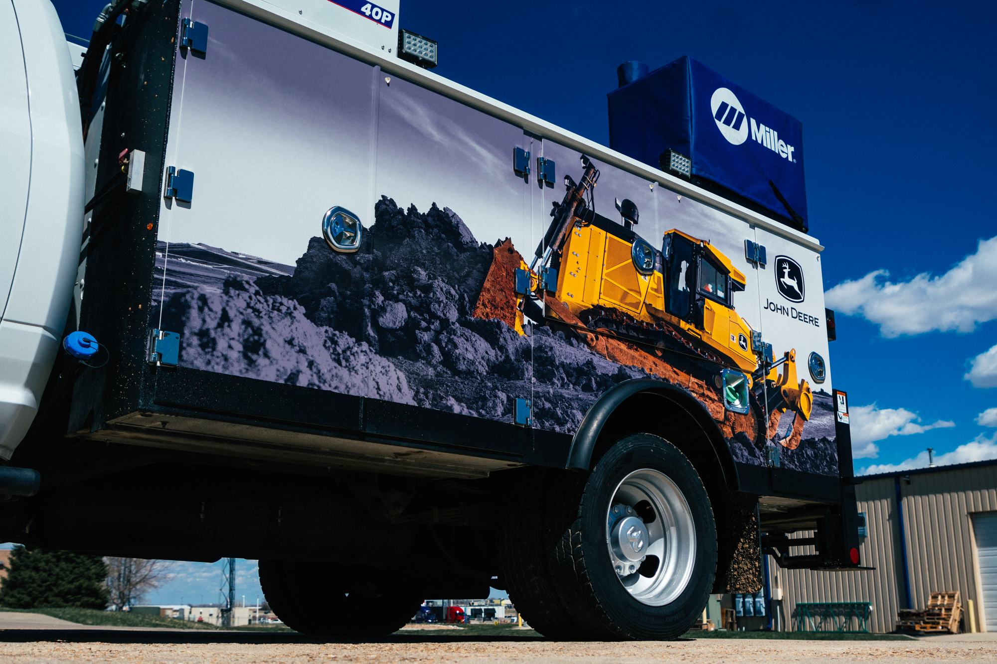 A partial vehicle wrap designed for 4Rivers in Fredrick, Colorado. The wrap depicts a powerful yellow John Deere bulldozer working on rocky terrain. The image is applied to the side of a white utility truck with the John Deere logo and branding on the vehicle's rear panel.